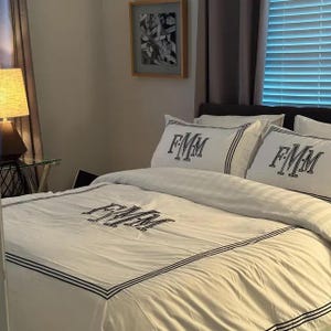 Puede incluir: Juego de ropa de cama blanco con ribete negro y las letras bordadas "FMM" en la funda n&oacute;rdica y las fundas de almohada. La ropa de cama est&aacute; sobre una cama en un dormitorio con una l&aacute;mpara y un cuadro enmarcado.