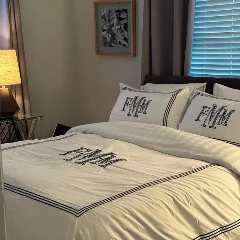 Puede incluir: Juego de ropa de cama blanco con ribete negro y las letras bordadas "FMM" en la funda n&oacute;rdica y las fundas de almohada. La ropa de cama est&aacute; sobre una cama en un dormitorio con una l&aacute;mpara y un cuadro enmarcado.