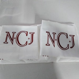 May include: White linens with the initials "NCJ" in burgundy. The initials are printed in a serif font. The linens are folded and stacked. The background is white.