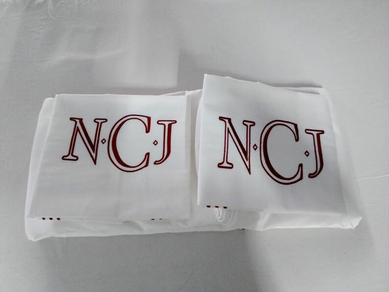 May include: White linens with the initials "NCJ" in burgundy. The initials are printed in a serif font. The linens are folded and stacked. The background is white.