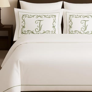 May include: White bedding set with green embroidered details. The set includes a duvet cover and two pillowcases. Each pillowcase features a green embroidered frame and the letter "J". The bed is made up and ready for use.