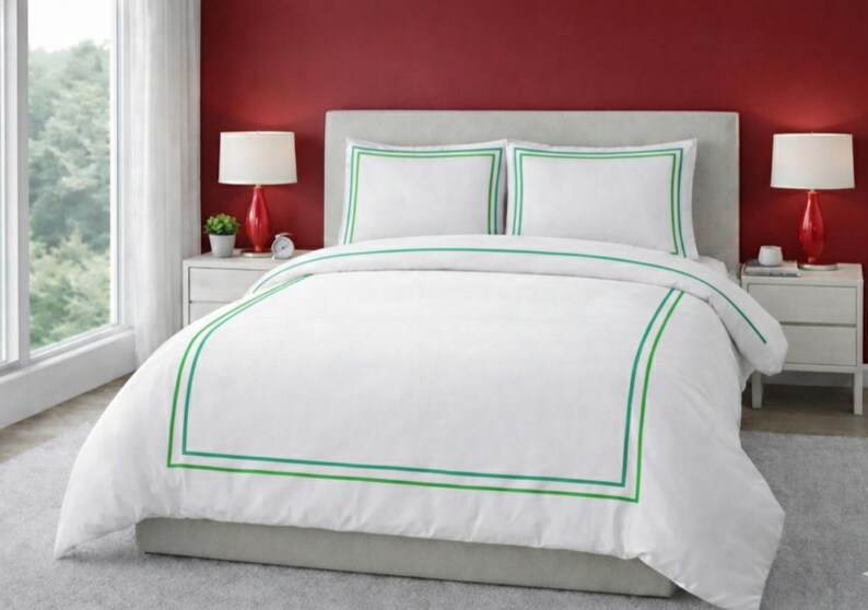 May include: White bedding set with green trim. The set includes a duvet cover and two pillowcases. The bedding is on a bed with a gray headboard. Red lamps sit on white nightstands on either side of the bed.