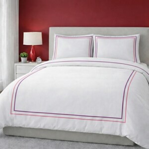 May include: White bedding set with a duvet cover and two pillowcases. The duvet and pillowcases have a purple and pink border. The bed is in a bedroom with red walls and two red lamps.