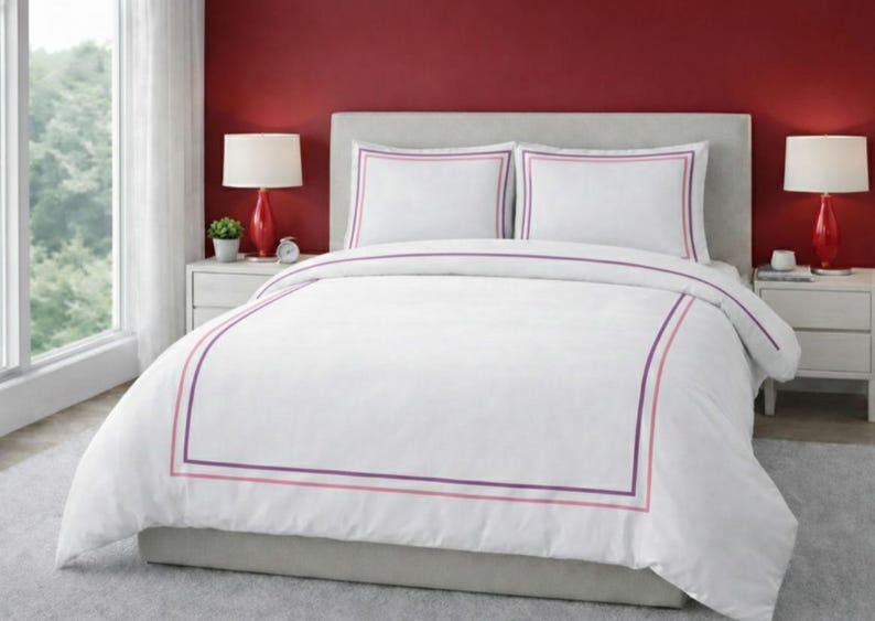 May include: White bedding set with a duvet cover and two pillowcases. The duvet and pillowcases have a purple and pink border. The bed is in a bedroom with red walls and two red lamps.