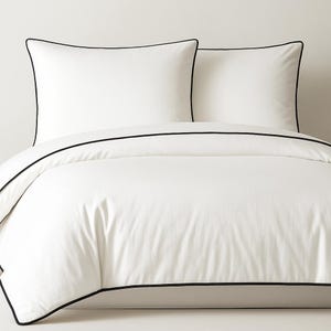 Puede incluir: Una funda nórdica blanca y dos fundas de almohada a juego con ribete negro. El juego de cama está hecho de una tela suave y lisa. Las fundas de almohada son cuadradas y la funda nórdica está doblada.