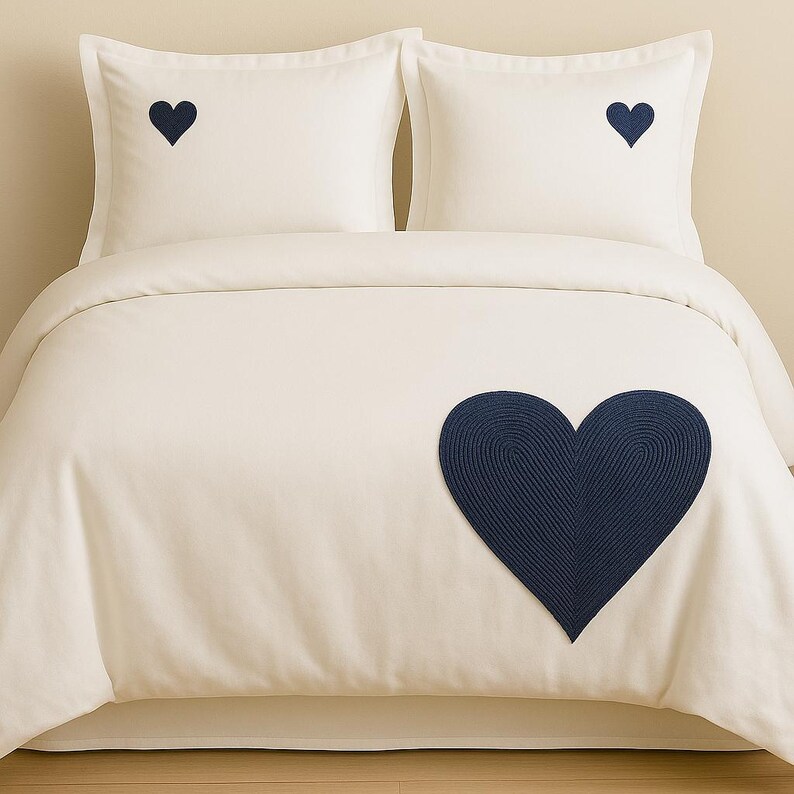 May include: A cream-colored bedding set consisting of a duvet cover and two pillowcases. Each pillowcase features a small, dark navy blue embroidered heart. The duvet cover displays a large, textured dark navy blue heart appliqu&eacute;.