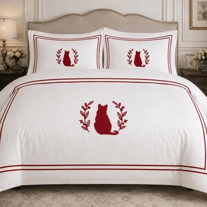 Pu&ograve; includere: Un set di biancheria da letto bianco composto da un copripiumino e due federe con la sagoma di un gatto ricamata in rosso, centrata tra due rami frondosi. Il design presenta una doppia cornice rossa sul copripiumino e una singola sulle federe.