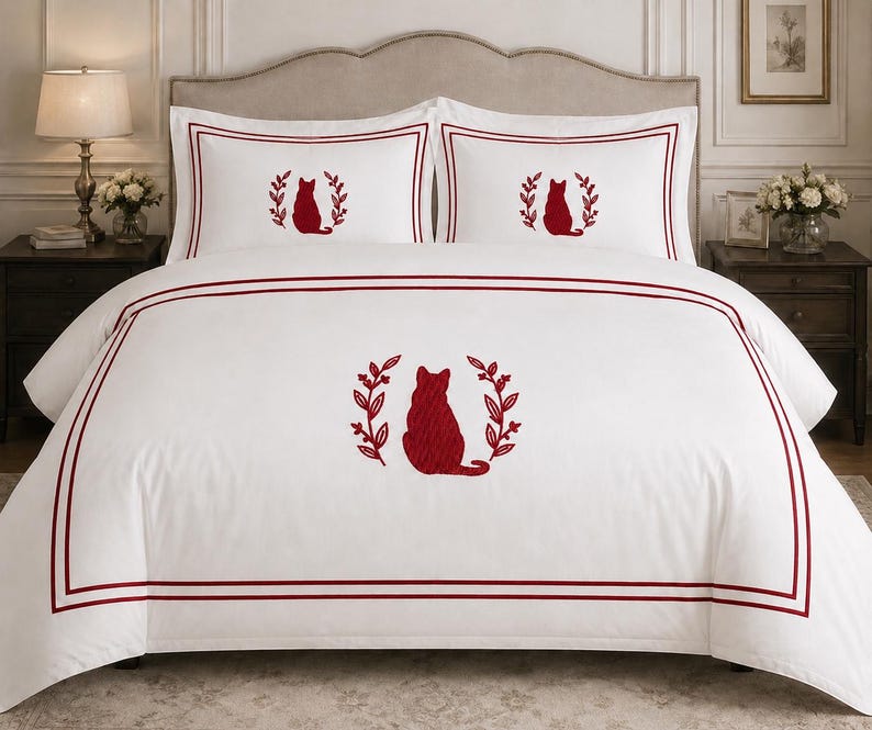 Pu&ograve; includere: Un set di biancheria da letto bianco composto da un copripiumino e due federe con la sagoma di un gatto ricamata in rosso, centrata tra due rami frondosi. Il design presenta una doppia cornice rossa sul copripiumino e una singola sulle federe.