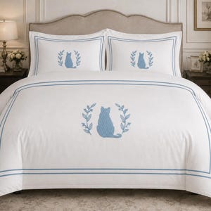 Pu&ograve; includere: Un set di biancheria da letto bianco composto da un copripiumino e due federe, ciascuno caratterizzato da una sagoma di gatto ricamata in blu all'interno di una corona di foglie blu e incorniciato da un doppio bordo rettangolare blu.