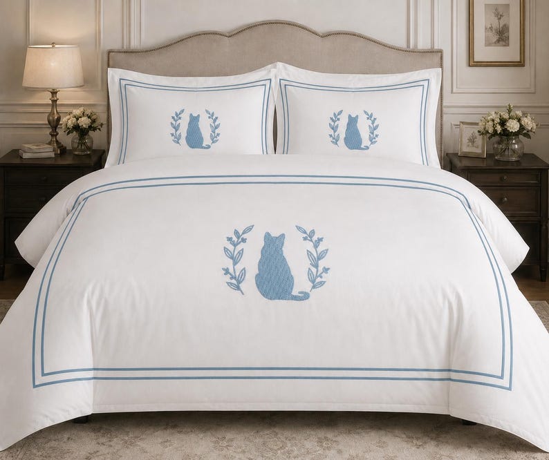 Pu&ograve; includere: Un set di biancheria da letto bianco composto da un copripiumino e due federe, ciascuno caratterizzato da una sagoma di gatto ricamata in blu all'interno di una corona di foglie blu e incorniciato da un doppio bordo rettangolare blu.