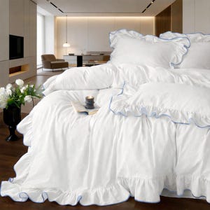 May include: White bedding set with ruffled edges and a light blue trim. The set includes a duvet cover and pillowcases. A book and candle sit on the bed. The room has a modern design with a dark wood floor.