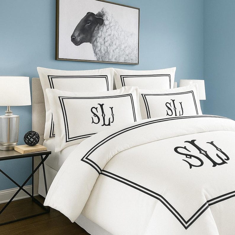 May include: White bedding set with black trim and monogrammed initials. The set includes a duvet cover and pillowcases. A framed black and white sheep portrait hangs above the bed. The room has light blue walls.