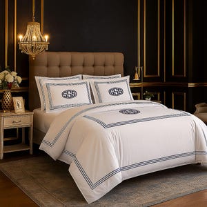Puede incluir: Juego de cama blanco con detalles bordados en azul marino. El juego incluye una funda n&oacute;rdica y dos fundas de almohada, cada una con un dise&ntilde;o de monograma. La cama est&aacute; en un dormitorio lujoso con una l&aacute;mpara de ara&ntilde;a y detalles dorados.