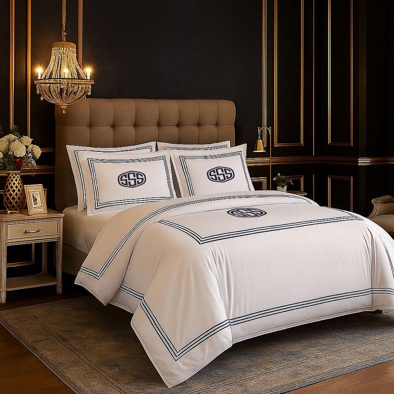 Puede incluir: Juego de cama blanco con detalles bordados en azul marino. El juego incluye una funda n&oacute;rdica y dos fundas de almohada, cada una con un dise&ntilde;o de monograma. La cama est&aacute; en un dormitorio lujoso con una l&aacute;mpara de ara&ntilde;a y detalles dorados.