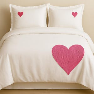 May include: A cream-colored bedding set consisting of a duvet cover and two pillowcases. Each pillowcase features a small, embroidered pink heart. The duvet cover is adorned with a large, textured pink heart made of coiled fabric.