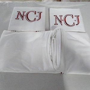 May include: White bed linen set featuring embroidered initials "NCJ" in burgundy on the pillowcases. The set includes a fitted sheet, a flat sheet, and two pillowcases, all neatly folded.