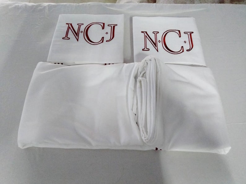 May include: White bed linen set featuring embroidered initials "NCJ" in burgundy on the pillowcases. The set includes a fitted sheet, a flat sheet, and two pillowcases, all neatly folded.