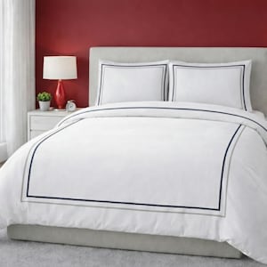 May include: White bedding set with a duvet cover and two pillowcases. The duvet cover and pillowcases have a navy blue and gray border. The bed is in a bedroom with red walls and two red lamps.