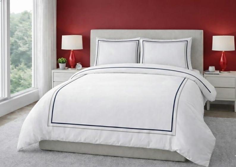 May include: White bedding set with a duvet cover and two pillowcases. The duvet cover and pillowcases have a navy blue and gray border. The bed is in a bedroom with red walls and two red lamps.