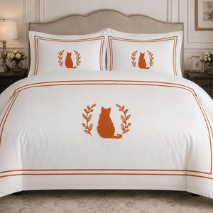 Pu&ograve; includere: Un set di biancheria da letto bianco composto da un copripiumino e due federe, ciascuno con una silhouette di gatto ricamata in arancione bruciato all'interno di una corona di foglie. Il tessuto presenta doppie linee di bordo arancioni.