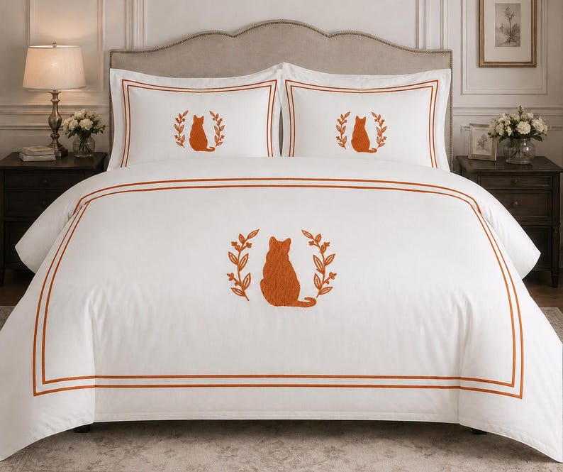 Pu&ograve; includere: Un set di biancheria da letto bianco composto da un copripiumino e due federe, ciascuno con una silhouette di gatto ricamata in arancione bruciato all'interno di una corona di foglie. Il tessuto presenta doppie linee di bordo arancioni.