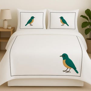 Puede incluir: Un juego de cama blanco que consta de una funda n&oacute;rdica y dos fundas de almohada, cada una con un borde rectangular negro fino y una ilustraci&oacute;n gr&aacute;fica de un p&aacute;jaro verde azulado y amarillo situado en el lado derecho del edred&oacute;n.