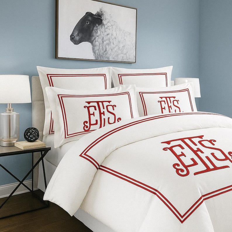 May include: White bedding set with red trim and embroidered initials. The set includes pillowcases and a duvet cover. A framed black and white sheep portrait hangs above the bed, creating a classic and elegant bedroom aesthetic.