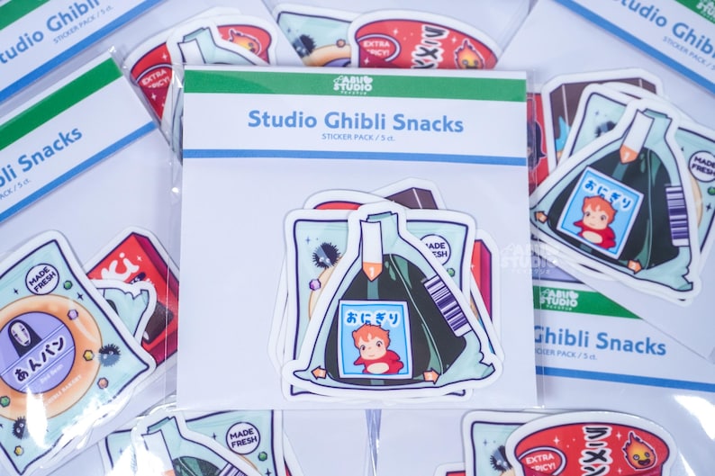 May include: A pack of Studio Ghibli Snacks stickers featuring various food-themed designs. The central pack displays an onigiri sticker with a cartoon character. Other stickers include ramen and other Japanese snacks.