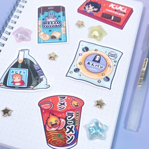 May include: A collection of anime-themed stickers on a spiral notebook. Stickers include a Hello Totoro snack, a Kiki's Delivery Service chocolate bar, and a cup of ramen. Also included are star-shaped embellishments and pens.