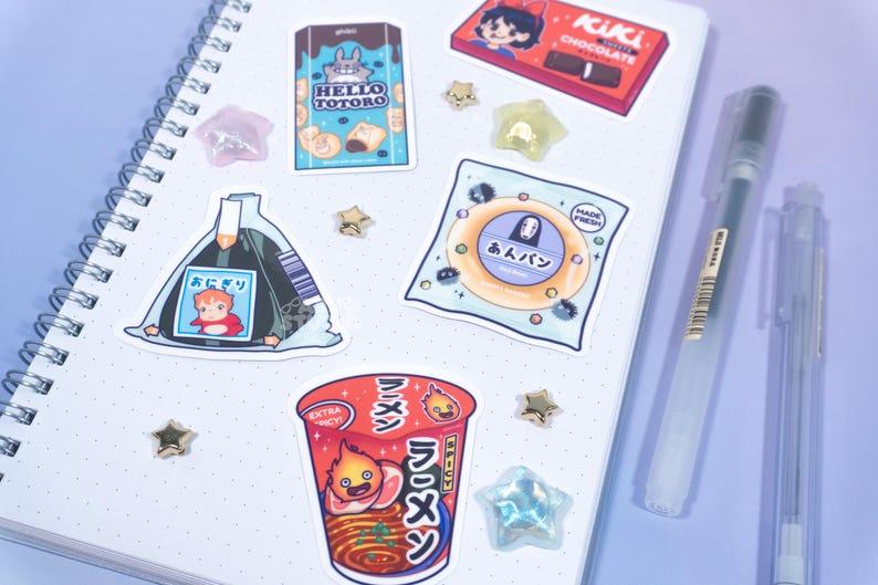 May include: A collection of anime-themed stickers on a spiral notebook. Stickers include a Hello Totoro snack, a Kiki's Delivery Service chocolate bar, and a cup of ramen. Also included are star-shaped embellishments and pens.