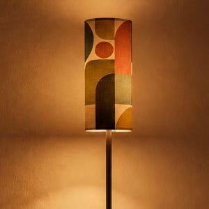 May include: A tall floor lamp with a slim black metal pole and a cylindrical lampshade featuring a geometric pattern in shades of olive green, burnt orange, and cream. The lamp is shown both unlit and illuminated with a warm, soft glow.