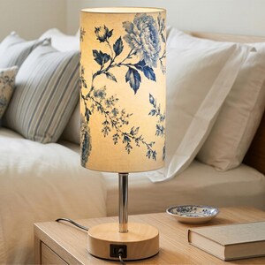 May include: A cylindrical table lamp featuring a cream-colored fabric shade with a blue floral pattern. The lamp has a polished chrome stem and a round light-colored wooden base that includes a built-in USB port and a black power cord.