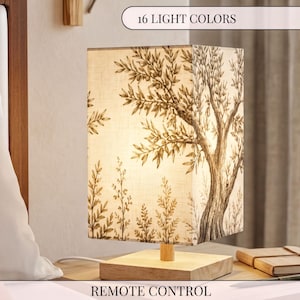 May include: A rectangular table lamp with a light-colored wooden base and a fabric shade featuring a dark brown ink-style illustration of an olive tree and foliage. The lamp includes a remote control and offers 16 light colors for illumination.