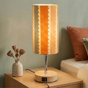May include: A table lamp with a cylindrical shade featuring vertical stripes in muted green, orange, and cream with scalloped edges. The lamp has a polished chrome stem and a round, light-colored wooden base with a built-in USB charging port.