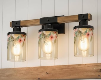 May include: A three-light vanity fixture featuring a horizontal wooden beam with black metal accents. Three cylindrical glass shades are decorated with a delicate pink floral and green leaf pattern, each housing a warm glowing light bulb.