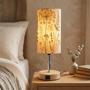 May include: A cylindrical table lamp with a light-colored wooden base and a chrome stem. The lampshade features a botanical print of dried flowers, ferns, and grasses in shades of brown and beige. The lamp emits a warm glow from within.