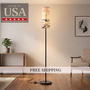 May include: A tall, black floor lamp with a cylindrical lampshade featuring a coastal scene with a sailboat. The lamp is positioned in a room with a wooden floor, a neutral-toned chair, and a framed geometric artwork. The text "MADE IN USA" is visible.
