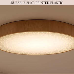 May include: A round flush mount ceiling light fixture with a light wood grain frame and a textured, translucent white diffuser. The text DURABLE FLAT-PRINTED PLASTIC is visible at the top. The light emits a warm, soft glow from the center.