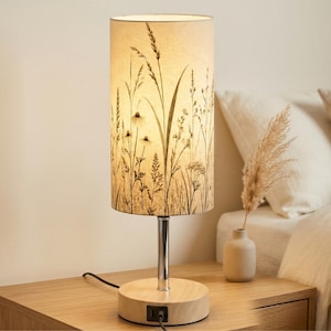 May include: A cylindrical table lamp with a light-colored fabric shade featuring a black botanical print of grasses and wildflowers. The lamp has a polished chrome metal stem and a round light-colored wooden base with a built-in USB charging port.
