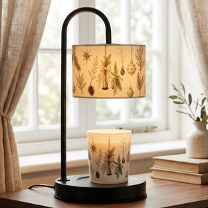 May include: A black metal candle warmer lamp with a curved arm and a cylindrical shade featuring a printed pattern of pine branches, pinecones, stars, and ornaments. A matching glass candle jar sits on the circular base beneath the lamp shade.