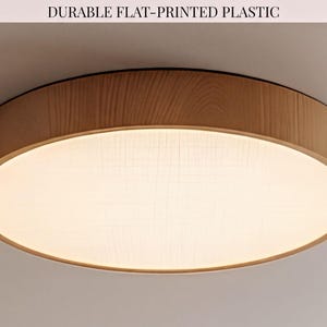 May include: A round flush mount ceiling light fixture with a light wood grain frame and a flat, off-white diffuser panel. The text DURABLE FLAT-PRINTED PLASTIC is visible at the top. The light emits a soft, warm glow across the circular surface.