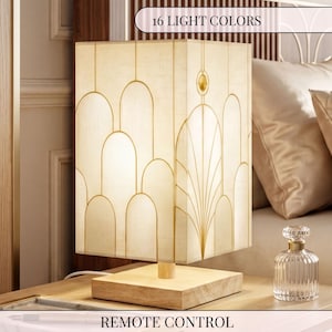 May include: A square table lamp with a light-colored fabric shade featuring gold-toned arched line patterns and a circular accent. The lamp has a light wood base and includes a remote control for 16 light colors. The design is geometric.