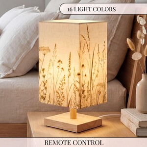 May include: A square table lamp with a light-colored fabric shade featuring a dark brown botanical print of wildflowers and grasses. The lamp has a light wood base and a short wooden stem. The text 16 LIGHT COLORS and REMOTE CONTROL is visible.