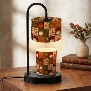 May include: A black metal candle warmer lamp with a curved arm and a cylindrical shade featuring a patchwork pattern of orange pumpkins, green leaves, and floral motifs. The base holds a matching candle jar with the same pumpkin and floral print.