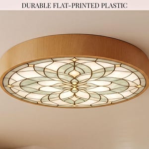 May include: A round ceiling light fixture with a light wood grain frame and a flat-printed plastic diffuser featuring a symmetrical, multi-petaled floral pattern in shades of white, cream, and soft green with dark outlines. Text reads: DURABLE FLAT-PRINTED PLASTIC.