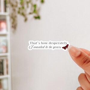Puede incluir: Una pegatina blanca con el texto "That's how desperately I wanted to be yours" y una pequeña ilustración de mariposa roja. La pegatina está sujeta por los dedos de una persona sobre un fondo blanco.