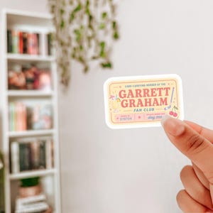 May include: A rectangular sticker with a cream background and a yellow border. The text reads "CARD-CARRYING MEMBER OF THE GARRETT GRAHAM FAN CLUB". It features a cocktail, a hockey stick, and two cherries in pink, blue, and yellow colours.