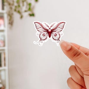 Könnte beinhalten: Ein roter und weißer Schmetterlingsaufkleber mit filigranen Details. Der Schmetterling hat eine zarte, fast durchscheinende Optik.