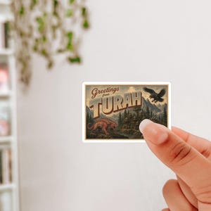May include: A sticker featuring a vintage postcard design with the text "Greetings from Turah." The illustration includes a dinosaur, an eagle, and a mountain landscape, with a sepia-toned color scheme.
