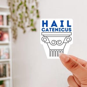 Op de afbeelding: Een witte sticker met de tekst "HAIL CATENICUS" in blauw en zwart. De sticker heeft een zwart-wit illustratie van een klassieke zuil. De sticker wordt door een hand vastgehouden.